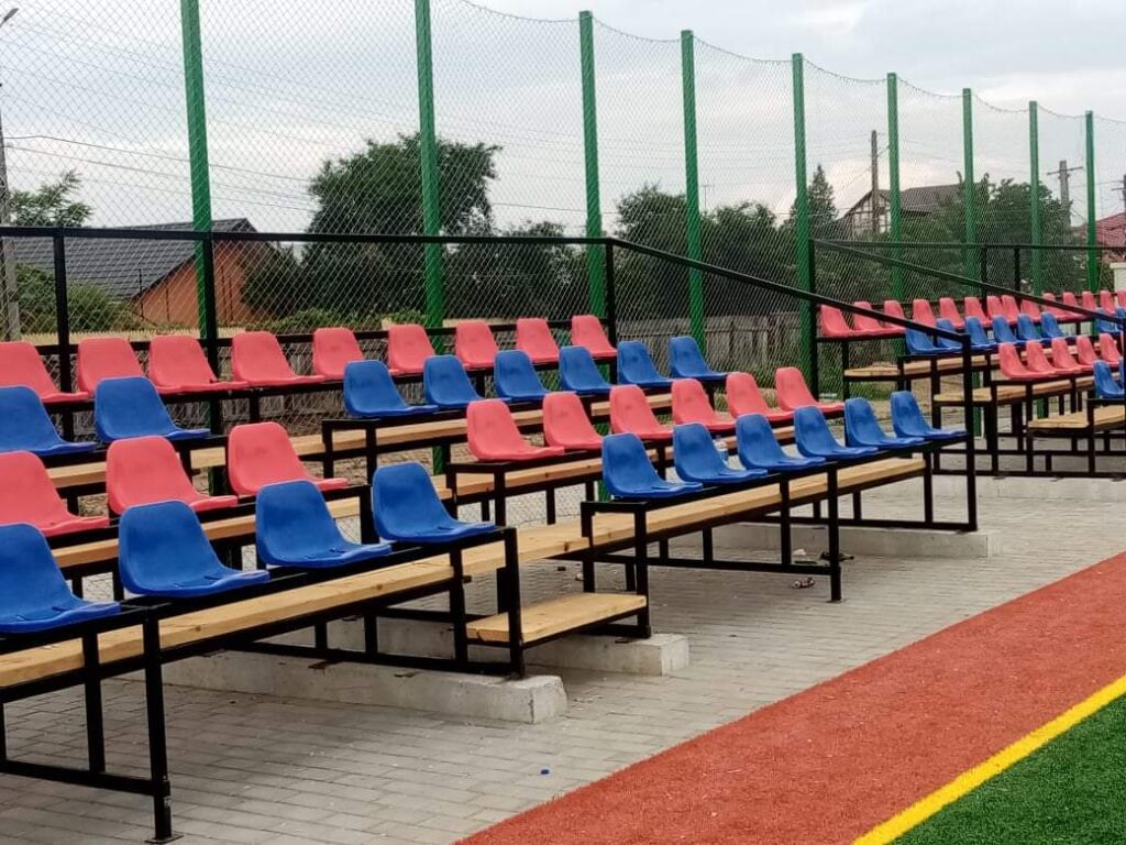 Tribună cu scaune lângă un teren de sport împrejmuit cu plasă verde de protecție
