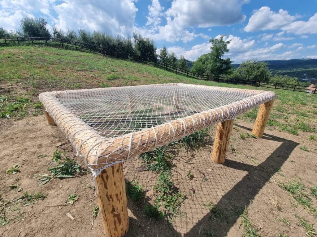 Pat din plasă albă montat pe o ramă din bușteni, pe o pajiște cu priveliște peste vale
