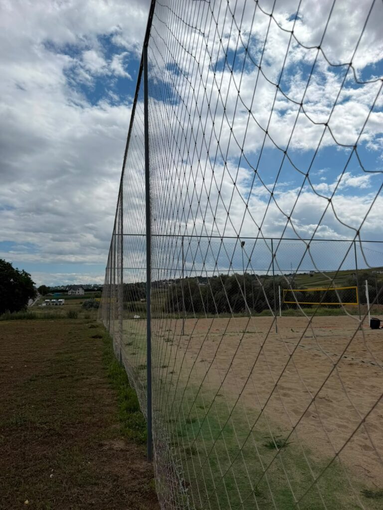 Plasă de împrejmuire înaltă pe stâlpi metalici lângă un teren de volei pe nisip, sub cer cu nori